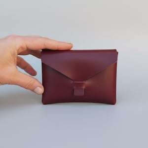 May include: A burgundy leather wallet with a flap closure. The wallet is rectangular and has a simple, minimalist design. The flap is secured with a small tab. The wallet is held by a hand.