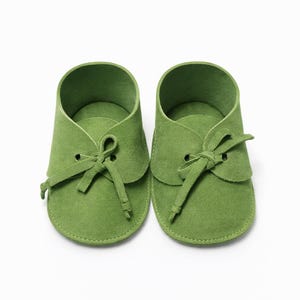 May include: A pair of green suede baby shoes. The shoes have a classic design with a rounded toe, a lace-up closure, and a soft, flexible sole. The shoes are a vibrant green color and are likely made of high-quality suede material.