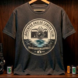 May include: Dark gray t-shirt with a circular graphic featuring a vintage camera design. The graphic includes the text "Järvenpää Analog Camera Club" and "Grain Over Megapixels Est. 2024". The shirt is hanging on a wooden hanger.