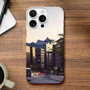 Könnte beinhalten: Eine Handyhülle mit einer Stadtlandschaft mit moderner Architektur und einer Straße mit Autos und einem Bus. Die Hülle hat einen hellen Hintergrund mit Blick auf Gebäude und eine Straßenszene.