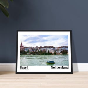 Könnte beinhalten: Schwarz gerahmter Druck mit einer malerischen Ansicht von Basel, Schweiz, mit der Skyline der Stadt entlang eines Flusses. Ein kleines Boot schwimmt auf dem Wasser. Der Druck enthält den Text "Basel" und "Switzerland".