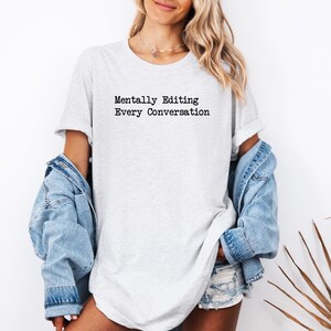 May include: A light gray short-sleeve t-shirt featuring the black text Mentally Editing Every Conversation printed on the chest. The shirt is paired with a light blue denim jacket and light blue denim shorts, creating a casual layered look.