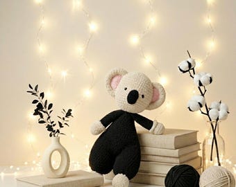 Könnte beinhalten: Ein gehäkeltes Koala-Stofftier mit cremefarbenem Kopf und schwarzem Körper, mit rosa Innenohren und einer schwarzen Nase. Der Koala sitzt auf einem Stapel Bücher neben einer weißen Vase mit dunklen Blättern, einem Baumwollzweig und Wollknäueln.