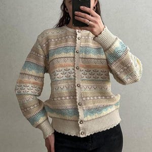 Op de afbeelding: Een button-down vest in crème, blauwe en perziktinten. Het gebreide kledingstuk heeft een patroon, een geschulpte zoom en geribde manchetten. Het heeft een ronde halslijn en lange mouwen. Een stijlvol gebreid kledingstuk.
