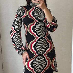 May include: A black mini dress with a geometric floral pattern in red, white, and tan. The dress has long sleeves with gathered cuffs and a high neckline. The pattern is repeated across the dress.