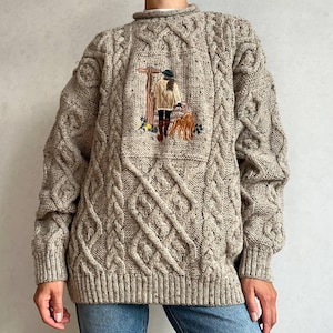 Op de afbeelding: Een beige kabeltrui met een gespikkelde textuur en een centraal geborduurd paneel. Het borduurwerk toont een figuur in een beige jas en donkere laarzen die een bruine hond uitlaat bij een houten wegwijzer met kleine gele bloemen.