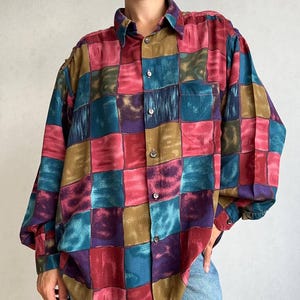 May include: A long-sleeve button-down shirt featuring a multi-colored checkered pattern in shades of red, teal, purple, and olive green. The shirt has a standard collar, a single chest pocket, and buttoned cuffs, with a loose, relaxed fit throughout.