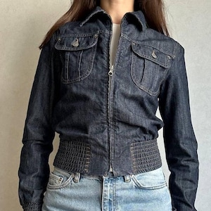Puede incluir: Una chaqueta de mezclilla azul oscuro con cremallera frontal completa, cuello clásico y dos bolsillos en el pecho con solapas abotonadas. La chaqueta incluye una cintura elástica ancha y acanalada, además de puños acanalados a juego.