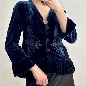 May include: A navy blue velvet jacket with long sleeves and a ruffled hem. The jacket features embroidered floral designs and three black buttons. The sleeves have a flared design. The jacket is a deep shade of blue.