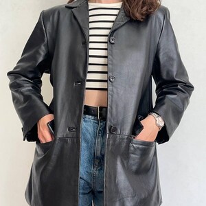 K&ouml;nnte beinhalten: Ein schwarzer Lederblazer mit Reverskragen, langen &Auml;rmeln und einer Knopfleiste mit drei Kn&ouml;pfen. Die Jacke verf&uuml;gt &uuml;ber zwei gro&szlig;e aufgesetzte Taschen an der H&uuml;fte und wird &uuml;ber einem wei&szlig;-schwarz horizontal gestreiften Oberteil getragen.