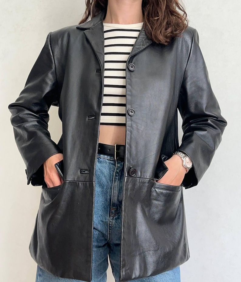 K&ouml;nnte beinhalten: Ein schwarzer Lederblazer mit Reverskragen, langen &Auml;rmeln und einer Knopfleiste mit drei Kn&ouml;pfen. Die Jacke verf&uuml;gt &uuml;ber zwei gro&szlig;e aufgesetzte Taschen an der H&uuml;fte und wird &uuml;ber einem wei&szlig;-schwarz horizontal gestreiften Oberteil getragen.