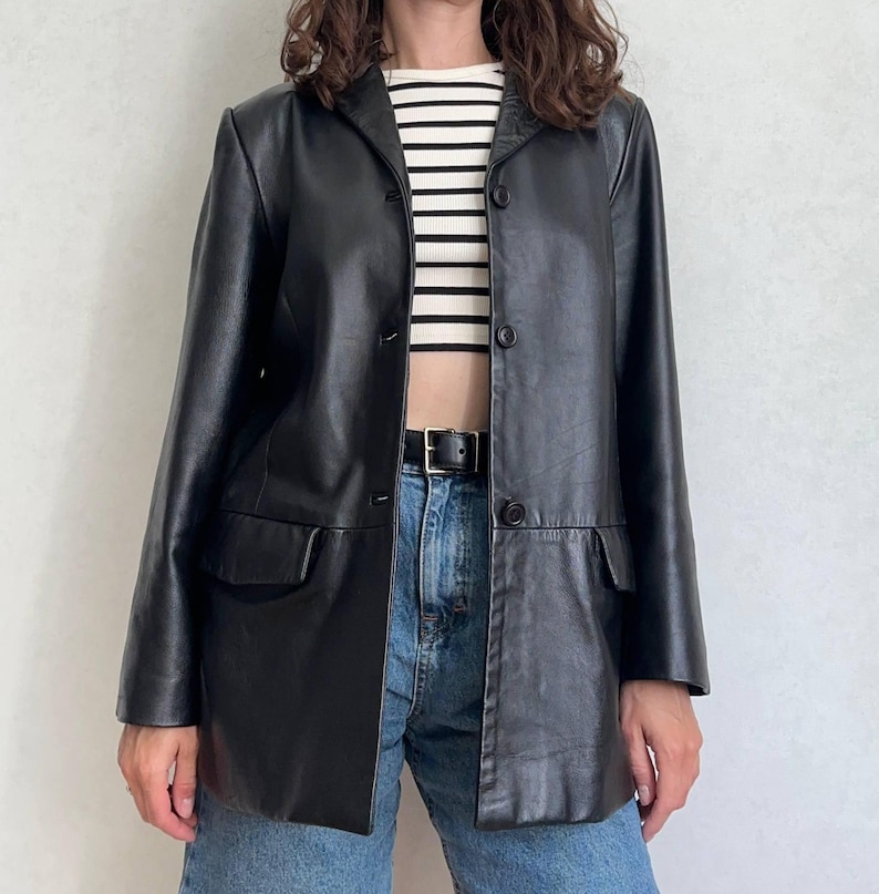 K&ouml;nnte beinhalten: Ein schwarzer Lederblazer mit Reverskragen, langen &Auml;rmeln und drei Kn&ouml;pfen auf der Vorderseite. Die Jacke verf&uuml;gt &uuml;ber zwei Pattentaschen und wird &uuml;ber einem bauchfreien Oberteil mit wei&szlig;en und schwarzen Querstreifen zu einer blauen Jeans getragen.