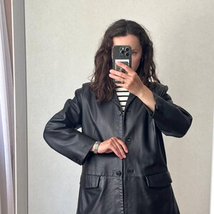 K&ouml;nnte beinhalten: Ein schwarzer Lederblazer mit steigendem Revers, einer dreikn&ouml;pfigen Vorderseite und zwei aufgesetzten Pattentaschen. Die Jacke hat lange &Auml;rmel mit gekn&ouml;pften Manschetten und eine strukturierte Passform, getragen &uuml;ber einem wei&szlig;-schwarz gestreiften Hemd.