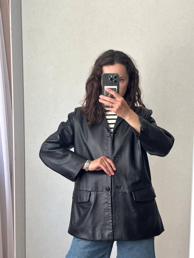 K&ouml;nnte beinhalten: Ein schwarzer Lederblazer mit steigendem Revers, einer dreikn&ouml;pfigen Vorderseite und zwei aufgesetzten Pattentaschen. Die Jacke hat lange &Auml;rmel mit gekn&ouml;pften Manschetten und eine strukturierte Passform, getragen &uuml;ber einem wei&szlig;-schwarz gestreiften Hemd.
