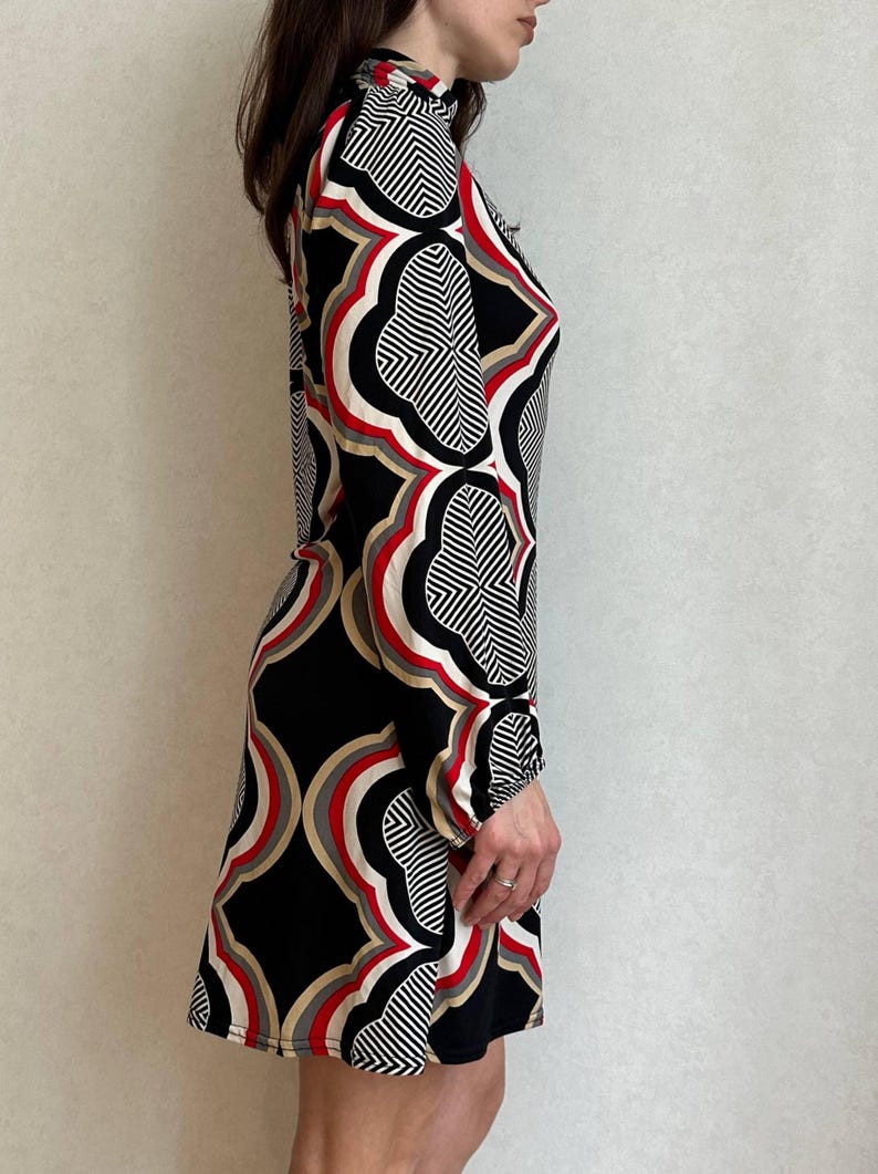 May include: A long-sleeved dress featuring a geometric pattern in black, white, red, and tan. The dress has a high neckline and a fitted silhouette, suitable for various occasions. The design incorporates repeating shapes and lines.