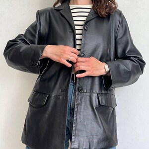 K&ouml;nnte beinhalten: Ein schwarzer Lederblazer mit fallendem Revers, langen &Auml;rmeln mit gekn&ouml;pften Manschetten und einer Knopfleiste vorne. Die Jacke verf&uuml;gt &uuml;ber zwei Pattentaschen auf Taillenh&ouml;he und wird &uuml;ber einem wei&szlig;-schwarz horizontal gestreiften Oberteil getragen.