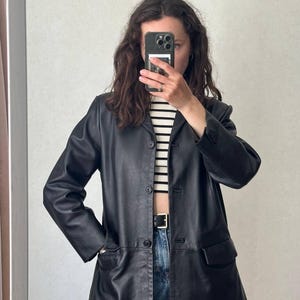 K&ouml;nnte beinhalten: Ein schwarzer Lederblazer mit Knopfleiste, langen &Auml;rmeln mit gekn&ouml;pften Manschetten und zwei Vordertaschen mit Klappen. Die Jacke wird &uuml;ber einem wei&szlig;-schwarz horizontal gestreiften Oberteil und einer blauen Jeans mit schwarzem G&uuml;rtel getragen.