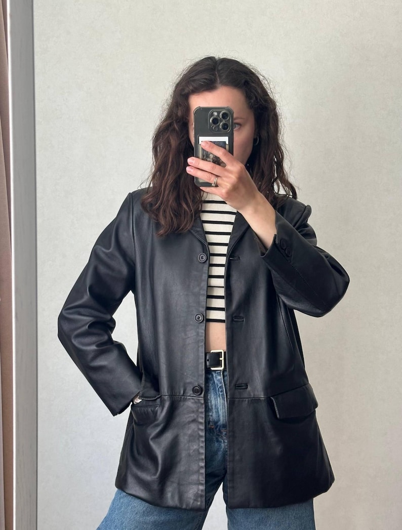 K&ouml;nnte beinhalten: Ein schwarzer Lederblazer mit Knopfleiste, langen &Auml;rmeln mit gekn&ouml;pften Manschetten und zwei Vordertaschen mit Klappen. Die Jacke wird &uuml;ber einem wei&szlig;-schwarz horizontal gestreiften Oberteil und einer blauen Jeans mit schwarzem G&uuml;rtel getragen.