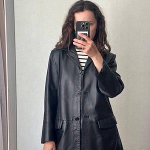 K&ouml;nnte beinhalten: Ein schwarzer Lederblazer mit fallendem Revers, einer dreikn&ouml;pfigen Knopfleiste und zwei Pattentaschen auf der Vorderseite. Die Jacke hat lange &Auml;rmel mit gekn&ouml;pften Manschetten und einen geraden Saum, getragen &uuml;ber einem schwarz-wei&szlig; gestreiften Oberteil.