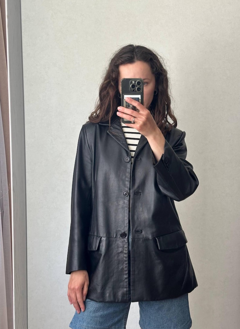 K&ouml;nnte beinhalten: Ein schwarzer Lederblazer mit fallendem Revers, einer dreikn&ouml;pfigen Knopfleiste und zwei Pattentaschen auf der Vorderseite. Die Jacke hat lange &Auml;rmel mit gekn&ouml;pften Manschetten und einen geraden Saum, getragen &uuml;ber einem schwarz-wei&szlig; gestreiften Oberteil.