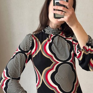 May include: A long-sleeved dress with a high neckline, featuring a bold geometric pattern in black, white, red, and beige. The dress has a fitted silhouette and a unique design, perfect for a vintage-inspired look.