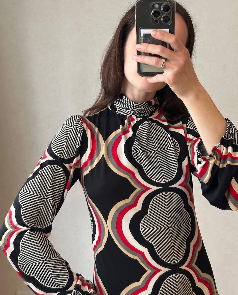 May include: A long-sleeved dress with a high neckline, featuring a bold geometric pattern in black, white, red, and beige. The dress has a fitted silhouette and a unique design, perfect for a vintage-inspired look.