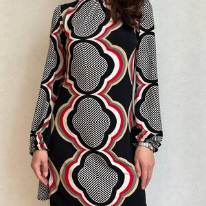 May include: A black, long-sleeved dress with a high neckline. The dress features a bold geometric pattern in white, red, and tan against a black background. The pattern is repeated throughout the dress, creating a striking visual effect.