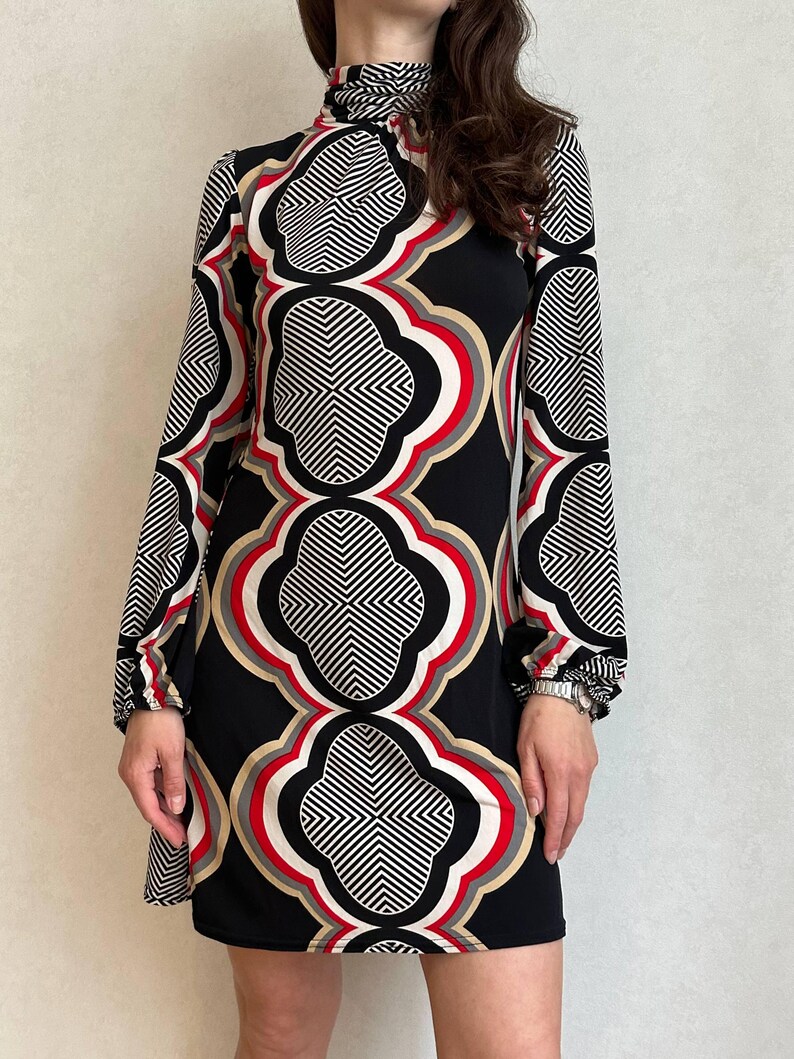 May include: A black, long-sleeved dress with a high neckline. The dress features a bold geometric pattern in white, red, and tan against a black background. The pattern is repeated throughout the dress, creating a striking visual effect.