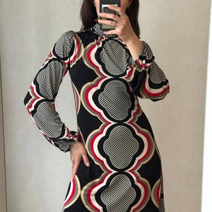 May include: A black dress with a geometric pattern in white, red, and gold. The dress has long sleeves with a ruffled cuff and a high neckline. The dress is knee-length.