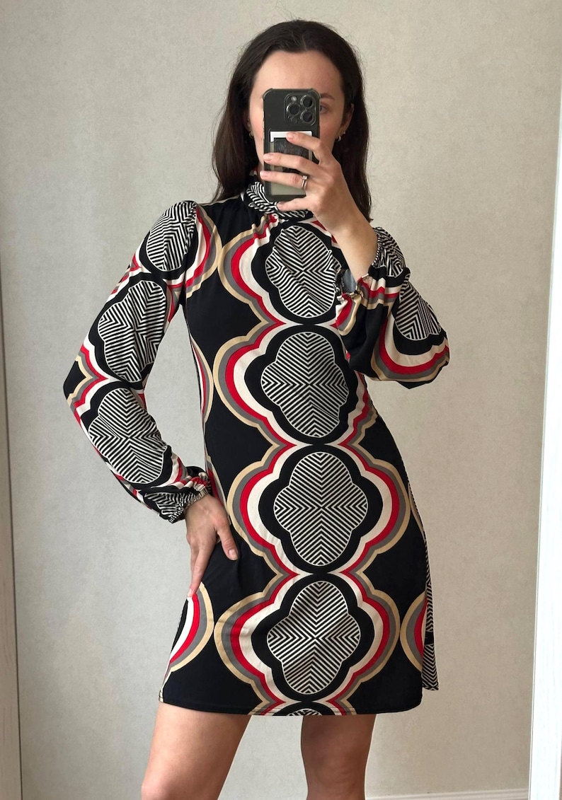 May include: A black dress with a geometric pattern in white, red, and gold. The dress has long sleeves with a ruffled cuff and a high neckline. The dress is knee-length.
