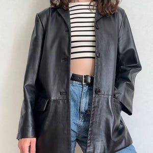 K&ouml;nnte beinhalten: Ein schwarzer Lederblazer mit steigendem Revers, drei Kn&ouml;pfen auf der Vorderseite und zwei Pattentaschen. Der Blazer wird &uuml;ber einem bauchfreien Oberteil mit wei&szlig;en und schwarzen Querstreifen und einer blauen Jeans mit schwarzem G&uuml;rtel getragen.