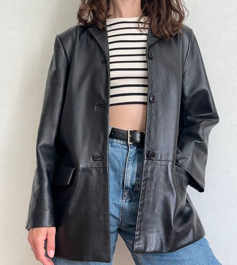 K&ouml;nnte beinhalten: Ein schwarzer Lederblazer mit steigendem Revers, drei Kn&ouml;pfen auf der Vorderseite und zwei Pattentaschen. Der Blazer wird &uuml;ber einem bauchfreien Oberteil mit wei&szlig;en und schwarzen Querstreifen und einer blauen Jeans mit schwarzem G&uuml;rtel getragen.