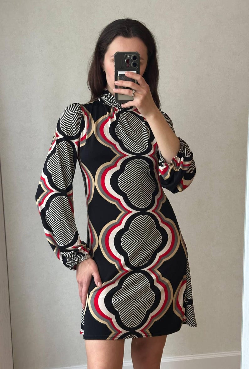 May include: A black mini dress with a geometric floral pattern in red, white, and tan. The dress has long sleeves with gathered cuffs and a high neckline. The pattern is repeated across the dress.