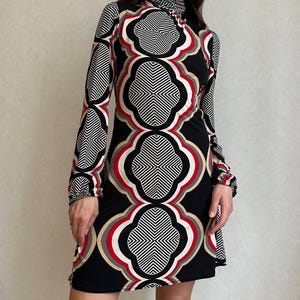 May include: A long-sleeved, black dress with a high neckline and a geometric pattern in white, red, and tan. The dress features a repeating quatrefoil design, creating a bold and eye-catching look. The dress is knee-length.