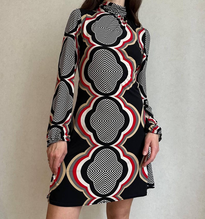 May include: A long-sleeved, black dress with a high neckline and a geometric pattern in white, red, and tan. The dress features a repeating quatrefoil design, creating a bold and eye-catching look. The dress is knee-length.