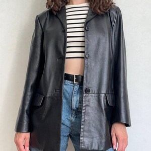 K&ouml;nnte beinhalten: Ein schwarzer Lederblazer mit Reverskragen, drei Kn&ouml;pfen auf der Vorderseite und zwei Pattentaschen. Der Blazer wird &uuml;ber einem gerippten Oberteil mit wei&szlig;en und schwarzen Querstreifen getragen, kombiniert mit einer blauen Jeans und einem schwarzen Lederg&uuml;rtel.
