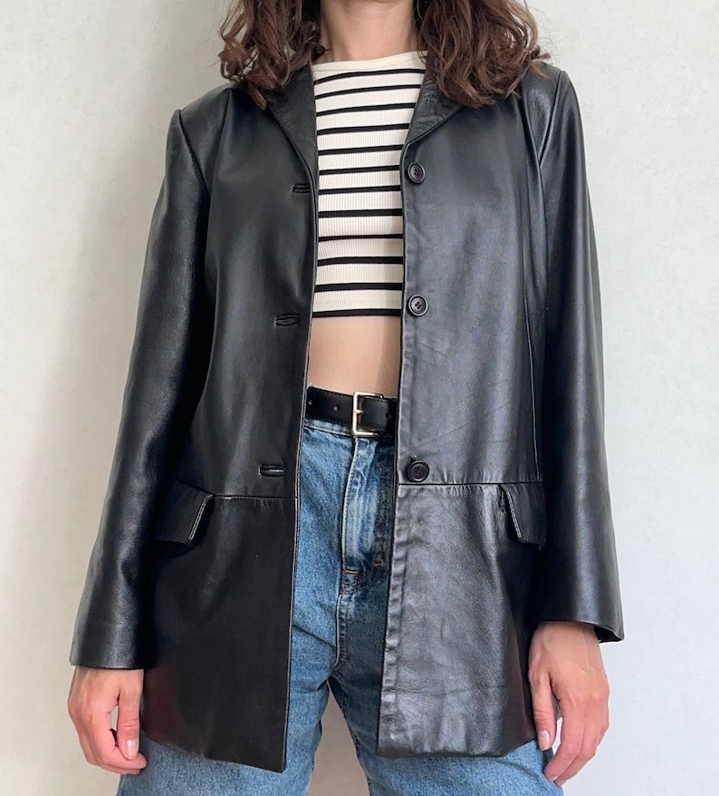 K&ouml;nnte beinhalten: Ein schwarzer Lederblazer mit Reverskragen, drei Kn&ouml;pfen auf der Vorderseite und zwei Pattentaschen. Der Blazer wird &uuml;ber einem gerippten Oberteil mit wei&szlig;en und schwarzen Querstreifen getragen, kombiniert mit einer blauen Jeans und einem schwarzen Lederg&uuml;rtel.