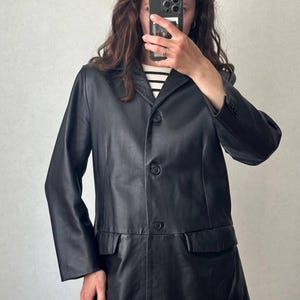 K&ouml;nnte beinhalten: Ein schwarzer Lederblazer mit steigendem Revers, drei Kn&ouml;pfen auf der Vorderseite und zwei Pattentaschen an der Taille. Die Jacke hat lange &Auml;rmel mit gekn&ouml;pften Manschetten und eine glatte Oberfl&auml;che, getragen &uuml;ber einem schwarz-wei&szlig; gestreiften Oberteil.
