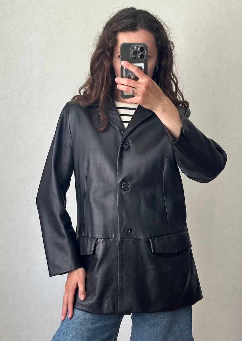 K&ouml;nnte beinhalten: Ein schwarzer Lederblazer mit steigendem Revers, drei Kn&ouml;pfen auf der Vorderseite und zwei Pattentaschen an der Taille. Die Jacke hat lange &Auml;rmel mit gekn&ouml;pften Manschetten und eine glatte Oberfl&auml;che, getragen &uuml;ber einem schwarz-wei&szlig; gestreiften Oberteil.