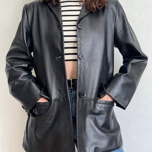 K&ouml;nnte beinhalten: Ein schwarzer Lederblazer mit Reverskragen, langen &Auml;rmeln und einer Knopfleiste mit drei Kn&ouml;pfen. Die Jacke verf&uuml;gt &uuml;ber zwei gro&szlig;e aufgesetzte Vordertaschen und wird &uuml;ber einem wei&szlig;-schwarz horizontal gestreiften Oberteil getragen.