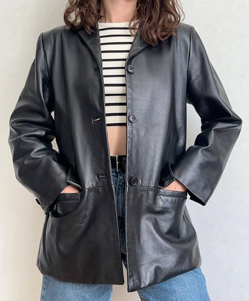 K&ouml;nnte beinhalten: Ein schwarzer Lederblazer mit Reverskragen, langen &Auml;rmeln und einer Knopfleiste mit drei Kn&ouml;pfen. Die Jacke verf&uuml;gt &uuml;ber zwei gro&szlig;e aufgesetzte Vordertaschen und wird &uuml;ber einem wei&szlig;-schwarz horizontal gestreiften Oberteil getragen.