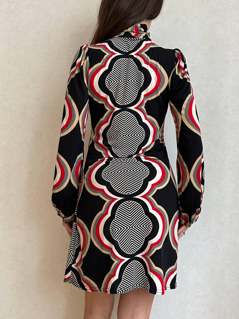 May include: A black dress with a geometric pattern in red, white, and tan. The long-sleeved dress has a high neckline and a fitted silhouette. The pattern is symmetrical and covers the entire dress.