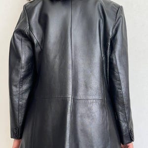 K&ouml;nnte beinhalten: Ein schwarzer Lederblazer mit einer Mittelnaht am R&uuml;cken, langen &Auml;rmeln mit zwei Kn&ouml;pfen an jedem &Auml;rmelabschluss und einer glatten Oberfl&auml;che. Die Jacke hat eine strukturierte Passform mit sichtbaren Paneelen am mittleren R&uuml;cken und den Schultern.