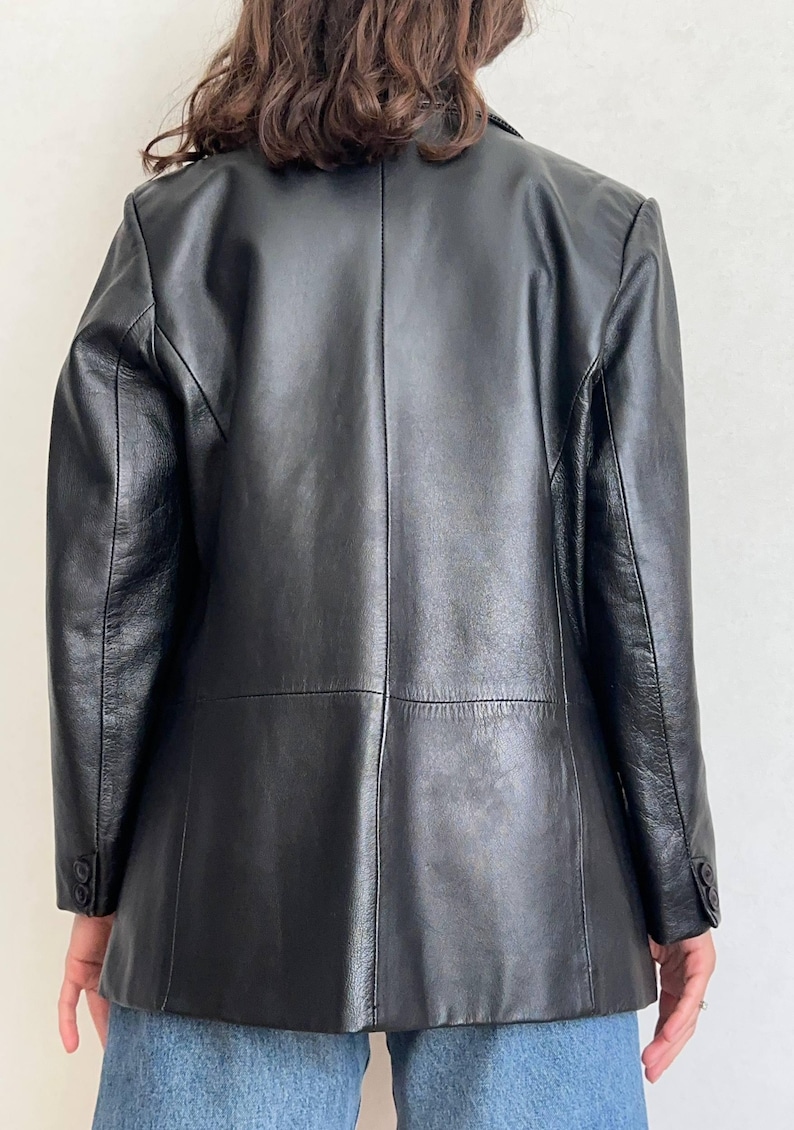 K&ouml;nnte beinhalten: Ein schwarzer Lederblazer mit einer Mittelnaht am R&uuml;cken, langen &Auml;rmeln mit zwei Kn&ouml;pfen an jedem &Auml;rmelabschluss und einer glatten Oberfl&auml;che. Die Jacke hat eine strukturierte Passform mit sichtbaren Paneelen am mittleren R&uuml;cken und den Schultern.