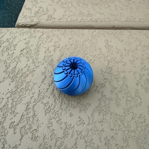 May include: A vibrant blue, spherical object with a ridged, floral-like design. The object is resting on a textured, light-colored surface. The design features curved lines radiating from the center.