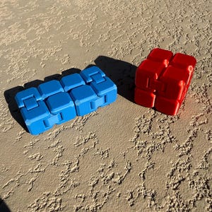 Pu&ograve; includere: Due giocattoli fidget: un cubo infinito blu rettangolare e un giocattolo fidget rosso a forma di cubo. I giocattoli sono fatti di plastica e sono progettati per essere manipolati. Lo sfondo &egrave; una superficie strutturata marrone chiaro.