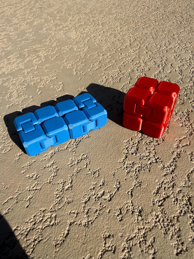 Pu&ograve; includere: Due giocattoli fidget: un cubo infinito blu rettangolare e un giocattolo fidget rosso a forma di cubo. I giocattoli sono fatti di plastica e sono progettati per essere manipolati. Lo sfondo &egrave; una superficie strutturata marrone chiaro.