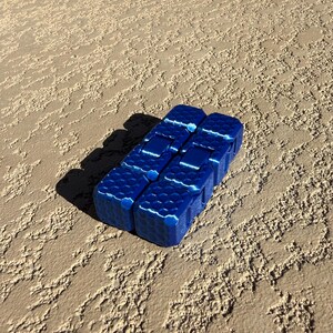 May include: A close-up of a blue fidget toy with a honeycomb pattern. The toy is made of several connected blocks. The blocks are a deep blue color with white accents. The toy is resting on a textured, tan surface.