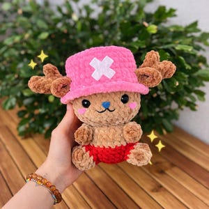 May include: A handmade plush toy of a cartoon character with a pink hat and brown antlers. The toy has a tan body, red shorts, and a blue nose. The background is a wooden surface with green foliage.