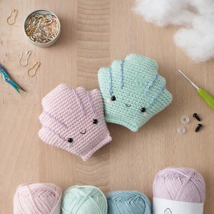 Könnte beinhalten: Zwei gehäkelte Spielzeuge in Muschelform, eines rosa und eines mintgrün, jeweils mit einem lächelnden Gesicht. Die Spielzeuge sind von Garn, Nähzubehör und Füllmaterial umgeben. Die Muscheln sind etwa 10 cm breit.